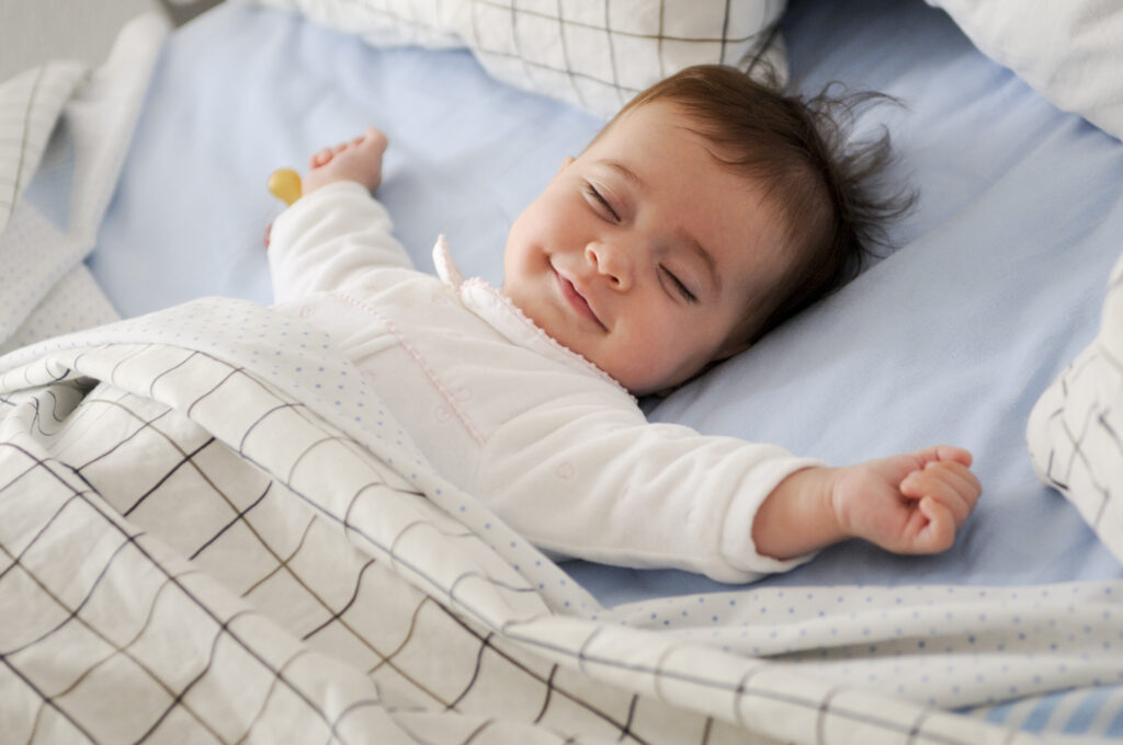 Bebê dormindo com um sorriso no rosto, com as mãozinhas fechadas e braços abertos