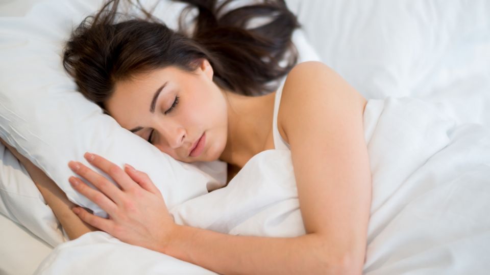 mulher dormindo de lado, com os braços embaixo do travesseiro, em uma cama com cobertas e fronhas brancas