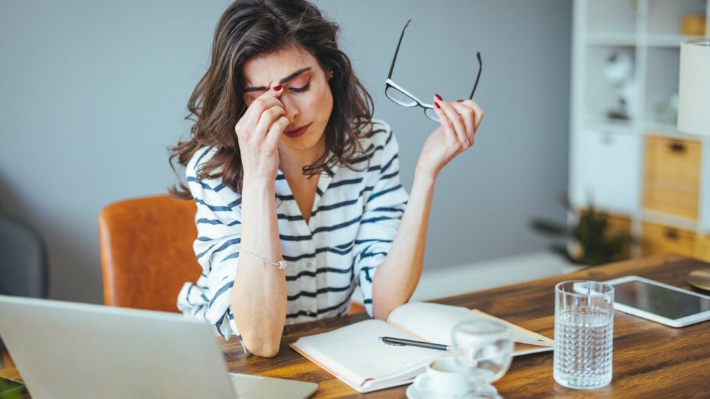 Mulher trabalhando de casa, numa mesa com notebook, um caderno, celular e um copo de água, estressada, segurando o óculos com uma das mãos e a outra segurando entre as sobrancelhas