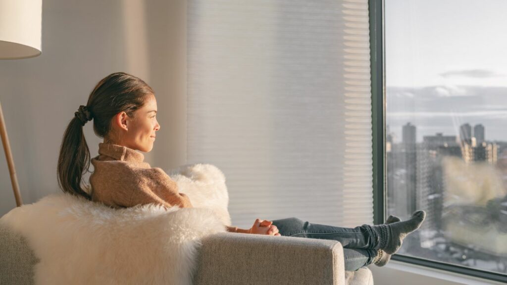 Mulher sentada em uma poltrona com um cobertor de pelo branco no encosto e pés esticados para frente em frente a janelas acústicas.
